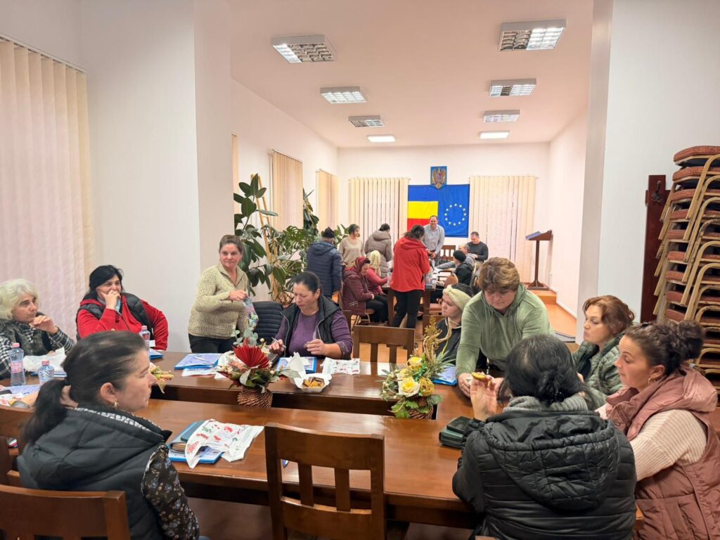 Participanți la un atelier comunitar, lucrând împreună la o activitate creativă, cu drapelele României și Uniunii Europene în fundal.